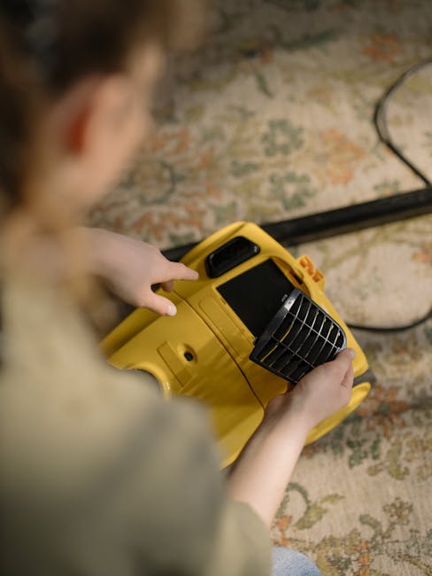 A person, likely using a commercial cleaning device, holds a yellow portable carpet or surface cleaning machine with a black vented front panel. The individual’s hands are positioned on the device, 