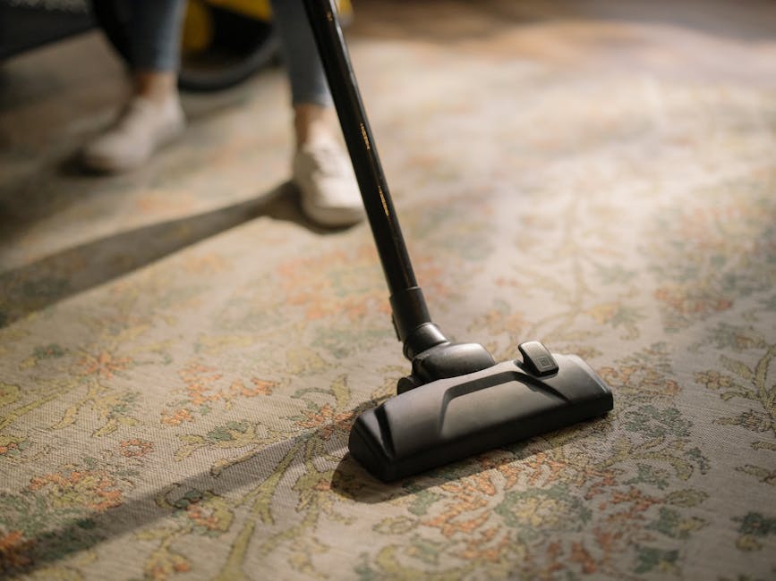 A woman with curly hair, dressed in a green jacket, blue jeans, and white sneakers, is engaged in deep surface cleaning in a living room. She is using a yellow vacuum cleaner with a hose attachment to